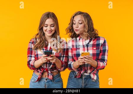 Image de deux jeunes filles belles portant des chemises à carreaux souriant et utilisant des téléphones cellulaires isolés sur fond jaune Banque D'Images