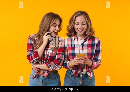 Image de deux jeunes filles belles portant des chemises à carreaux souriant et utilisant des téléphones cellulaires isolés sur fond jaune Banque D'Images