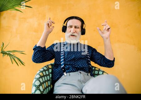 Photo en studio d'un homme âgé d'apparence soignée avec une barbe bien entretenue, assis sur la chaise et profitant de sa musique préférée dans des écouteurs. Isolé sur Banque D'Images