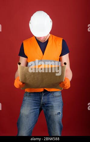 Le constructeur regarde le plan pour la nouvelle maison. Ingénieur, architecte, ouvrier, constructeur sur une face occupée tient le vieux plan en mains. Homme dans un casque, casque de sécurité tient le plan de construction, fond rouge. Concept d'ingénierie Banque D'Images