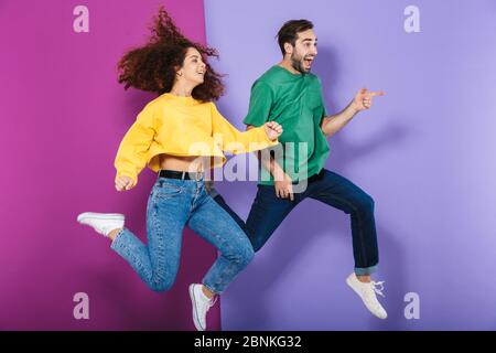 Portrait de l'homme et de la femme heureux de couple caucasien dans des vêtements colorés riant et courant ensemble isolé sur fond violet Banque D'Images