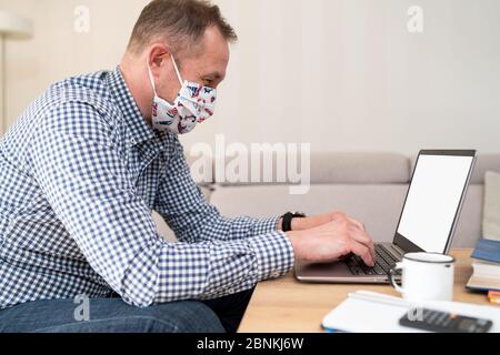 Portrait de beau jeune homme d'affaires avec masque médical chirurgical dans chemise à carreaux bleu sont assis et tapant le message sur l'ordinateur portable et regardant l'écran. Banque D'Images