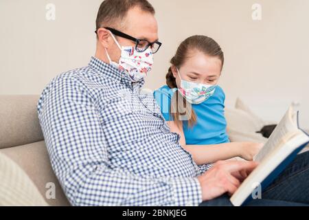 Papa dans le masque médical sur son visage lit livre avec enfant. Le père avec sa fille lit la Bible pendant une pandémie. Oncle aidant sa nièce avec les devoirs. Banque D'Images