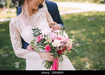 Gros plan du bouquet de pivoines, roses, eucalyptus d'une mariée dans des tons blanc-rose noués de rubans roses. La mariée tient un bouquet dans sa main dans un wh Banque D'Images