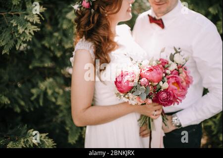 Bouquet de mariée gros plan, le bouquet se compose de pivoines rouges, roses rouges et blanches. La mariée tient le bouquet à côté du marié, vêtu d'un blanc Banque D'Images