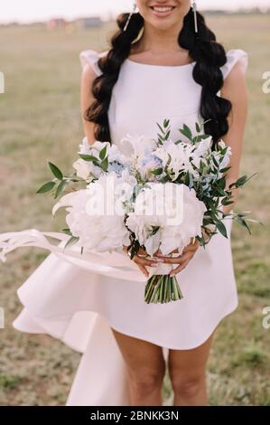 Gros plan du bouquet de pivoines d'une mariée, œillets dans des tons blancs et bleus. La mariée dans une robe blanche courte tient un bouquet dans ses mains. Le bouquet Banque D'Images