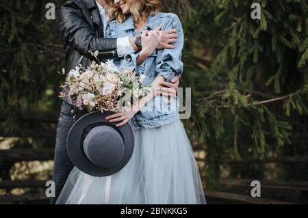 La mariée dans une veste en denim tient un chapeau et un bouquet dans les tons blanc et violet. La groom la déchagine de derrière Banque D'Images