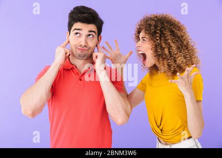 Portrait d'un jeune couple stressé arguant, homme se branchant les oreilles avec les doigts tandis que la femme hurlait isolé sur fond violet Banque D'Images