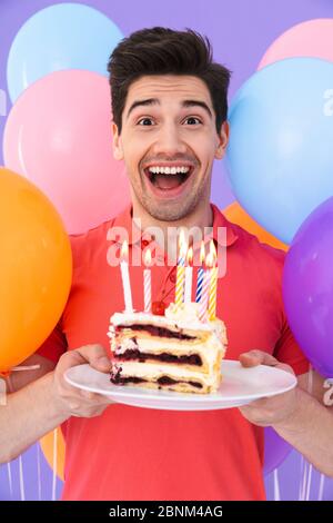 Image d'un bel homme joyeux célébrant son anniversaire avec des ballons d'air multicolores et un morceau de gâteau isolé sur fond violet Banque D'Images