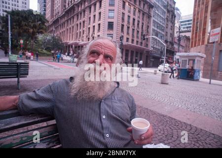 Sao Paulo, Brésil - 21 décembre 2015 : homme brésilien sans domicile avec une longue barbe blanche assis sur un banc pour boire un café et du lait Banque D'Images