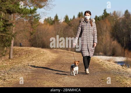 Une jeune femme en veste chaude, portant un masque facial de virus, marche son chien sur la route de campagne. Les masques sont obligatoires à l'extérieur de la maison pendant le coronavirus covid- Banque D'Images