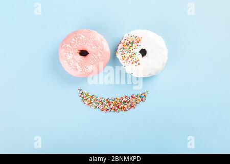 Beignet avec glaçage blanc et rose et garniture de pâtisserie au sucre multicolore sous forme de sourire sur fond bleu. Visage drôle de la nourriture. Vue de dessus Banque D'Images