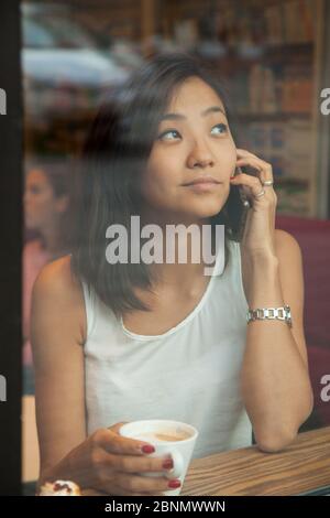 Belle jeune fille asiatique assise dans un café près de la fenêtre, boire du café et parler sur le téléphone.Focus sur une fille dans un café à travers le verre. Banque D'Images