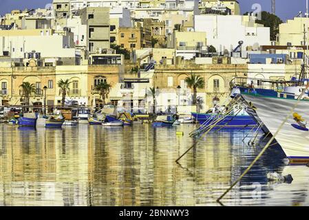 Ambiance pour le coucher du soleil dans le port de Marsaxlokk avec les bateaux de pêche typiques colorés, développement urbain dans le contexte de la montée Banque D'Images