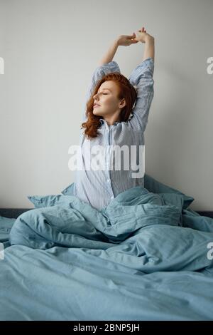 Une fille aux cheveux rouges en pyjama rayé est heureuse de se lever, assise sur le lit bleu Banque D'Images