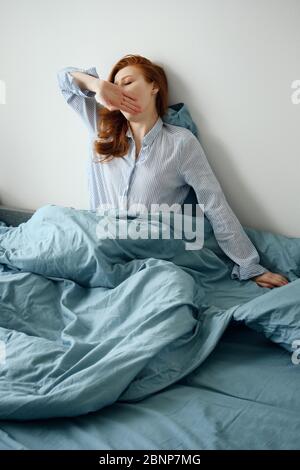 Une fille de pyjama à cheveux rouges et rayures couvre sa bouche avec sa main, assise dans un lit bleu Banque D'Images