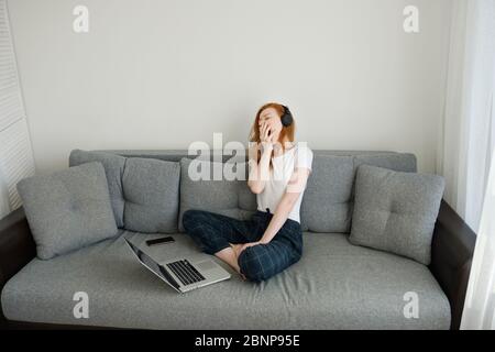 REDHEAD fille assise sur un canapé dans les vêtements et les écouteurs de la maison avec ordinateur portable et nawns largement, se cachant derrière la main, cadre d'en haut Banque D'Images