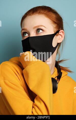 Vue de la tête. Une fille à cheveux rouges dans un sweat à capuche jaune se tient sur un fond bleu et sa main au masque noir regarde vers le haut Banque D'Images