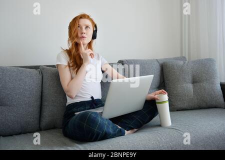 REDHEAD fille est assis sur un canapé dans des vêtements de maison avec un ordinateur portable sur les jambes croisées et regarde avec soin, avec un thermocup dans sa main Banque D'Images