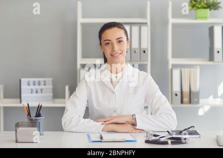 Médecin positif femme regardant la caméra souriant tout en étant assise à une table dans un bureau de clinique. Banque D'Images