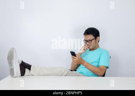 Jeune homme asiatique avec un visage heureux avec ce qu'il voit sur un téléphone portable avec une jambe sur la table. Indonésien portant une chemise bleue. Banque D'Images