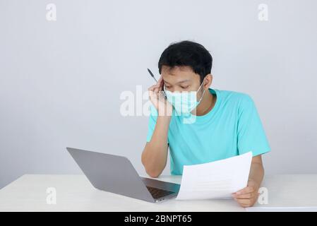 Le jeune homme asiatique portant un masque médical se sent malsain, fatigué et confus avec le travail dans les documents et les ordinateurs portables sur la table. Banque D'Images