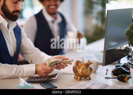 Gros plan sur un homme d'affaires comptant de l'argent gagné. Un partenaire d'affaires souriant à la réussite de ses collègues. La banque de porc comme symbole de fortune. Banque D'Images