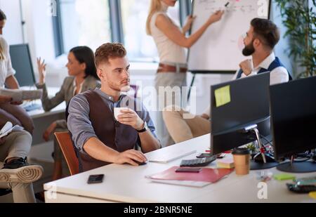 Jeune entrepreneur tenant une tasse de café tout en étant assis et travaillant dans le bureau plein de personnes Banque D'Images