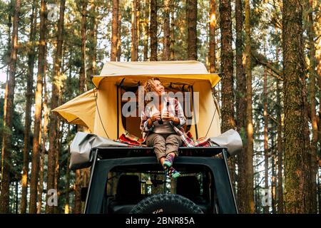 Voyage et style de vie wanderlust concept avec une femme heureuse solitaire adulte s'asseoir sur le toit tente véhicule de voiture avec forêt de bois en arrière-plan profiter de la nature et des vacances à l'extérieur Banque D'Images