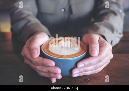 Gros plan de mains tenant une tasse bleue de café latte chaud avec art latte sur une table en bois dans le café Banque D'Images