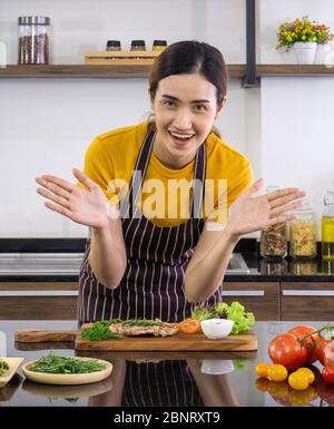 La femme de ménage vêtue d'un tablier, sourit et lève les deux mains pour offrir un délicieux steak sur le devant. Ambiance matinale dans une cuisine moderne Banque D'Images