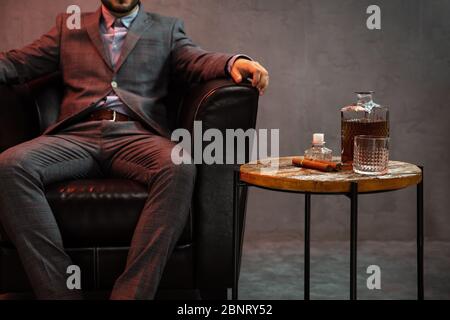 Gentleman est assis dans un fauteuil en cuir avec une carafe de whisky, un verre vide et un cigare sur la table. Banque D'Images