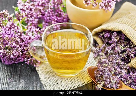 Thé à base de plantes origan dans une tasse de verre sur serviette en toile de jute, fleurs de marjolaine séchées dans un sac et une cuillère, fleurs fraîches dans le mortier et sur table sur une planche en bois Banque D'Images