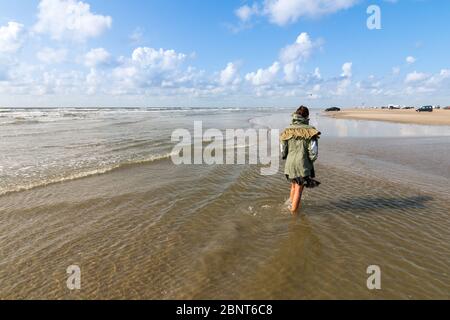 Une femme marche le long des eaux propres et transparentes de la plage de Romo, au Danemark, portant un manteau vert avec quelques voitures sur le sable en arrière-plan. Plage con Banque D'Images