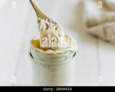 Prêt à préparer du pain de pâte de blé de démarrage goutte à goutte à partir d'une cuillère en bois. Pot en verre avec levain de départ 100 pour cent d'hydratation sur fond de bois blanc. Copier l'espace. Concept de pain maison Banque D'Images