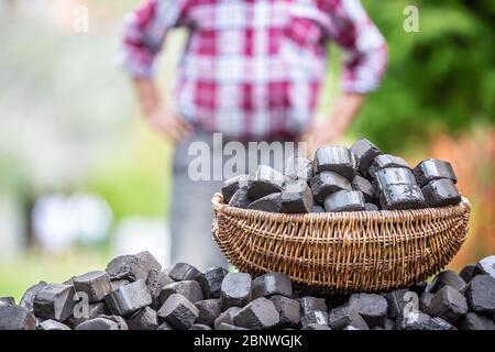 Panier rempli de briquettes de charbon sur une pile avec un homme debout en arrière-plan avec des bras sur sa taille. Banque D'Images