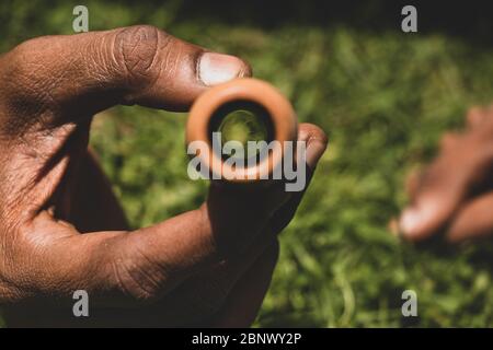 Main brune tenant et montrant la boule de réflexion verte de l'herbe derrière faite à l'intérieur d'un pipe indien de fumage (chillum), 5K Banque D'Images