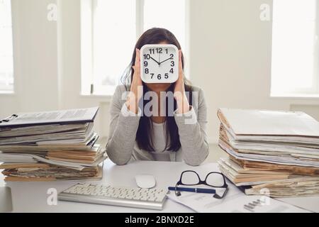 Femme d'affaires tenant un réveil devant son visage tout en étant assise à une table dans le bureau. Banque D'Images