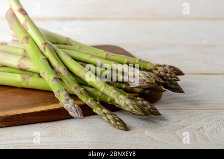 Tiges d'asperges vertes fraîches crues sur une planche à découper brune sur une table de cuisine en bois blanc. Ingrédient pour manger de la viande, régime végétarien et alimentaire cru. Banque D'Images