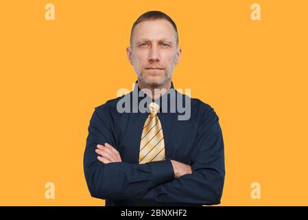 Homme dans une chemise bleu foncé et une cravate jaune vif et avec les bras croisés sur un fond orange. Banque D'Images