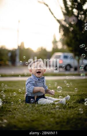 Bébé garçon portant un noeud papillon et regardant des bulles d'air assis dans le parc, saison du printemps à l'extérieur Banque D'Images