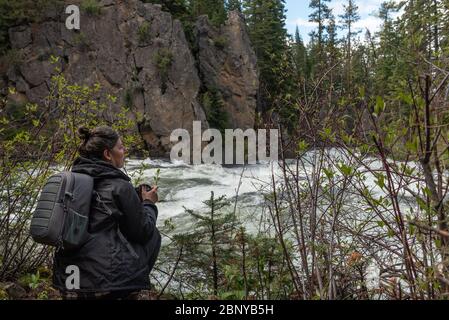 Femme d'âge moyen, portant un sac à dos gris et un appareil photo à la main, se préparer à photographier une rivière qui se précipite Banque D'Images