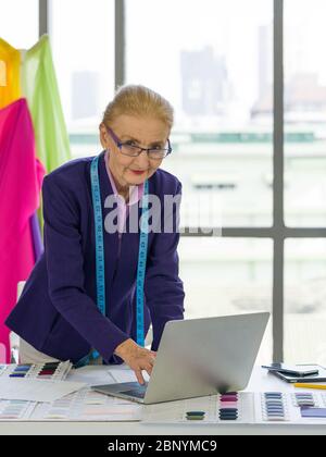 Une designer senior utilise son ordinateur portable pour rechercher des informations pour le design moderne. Un bureau rempli d'échantillons de tissus aux couleurs vives. Ambiance matinale Banque D'Images