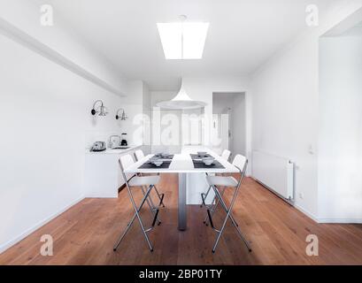 Nouvelle cuisine lumineuse et élégante avec armoires blanches. Intérieur moderne et spacieux avec parquet, table blanche, chaises industrielles et grande fenêtre au plafond. Banque D'Images