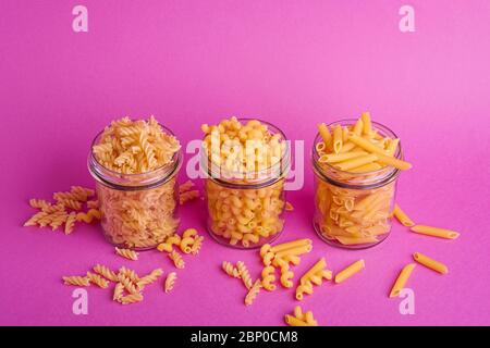 Trois pots en verre avec variété de pâtes de blé doré non cuites sur fond rose minimal, vue en angle Banque D'Images