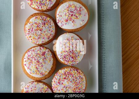 Gâteaux de fée glacés dans une rangée sur une longue plaque blanche placée sur un tissu bleu. Les gâteaux de fées sont couverts de saupoudrés de sucre multicolore. Banque D'Images