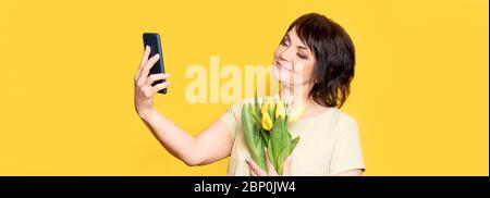 Belle vieille femme avec maquillage professionnel fumé et coiffure tenant des fleurs de tulipe sur le fond jaune. Prise de vidéos par le smartphone. Banque D'Images