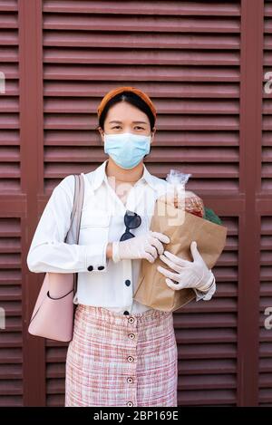 Femme asiatique tenant un sac à provisions tout en portant un masque et des gants pour prévenir la propagation du virus pendant la pandémie du coronavirus avec Copy Space ag Banque D'Images