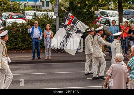 Les membres d'une association de vétérans de la marine portent l'étendard historique de la marine impériale allemande lors d'un défilé de fête de tir à Grevenbroich, en Allemagne – une fête traditionnelle allemande des tireurs. Le design, une croix de fer et un aigle sur un fond blanc, a été utilisé de 1903 à 1921. Deux passants font signe aux marcheurs Banque D'Images