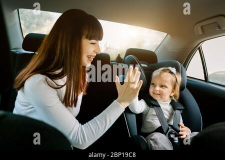 Jolie jeune femme mère prend soin de sa petite fille dans un siège de sécurité de voiture, assis ensemble, donnant élevé cinq l'un l'autre et s'amusant Banque D'Images